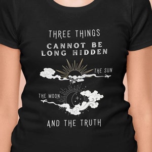 Puede incluir: Camiseta negra con un diseño gráfico blanco que presenta un sol, una luna y nubes. El texto "Three things cannot be long hidden" está por encima del gráfico y "The Sun", "The Moon", y "And the truth" están por debajo del gráfico.