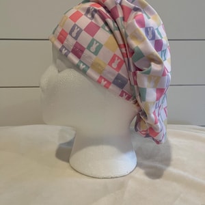 May include: A white scrub hat with a checkered pattern in pink, yellow, green, and purple. The pattern also includes white bunny silhouettes.