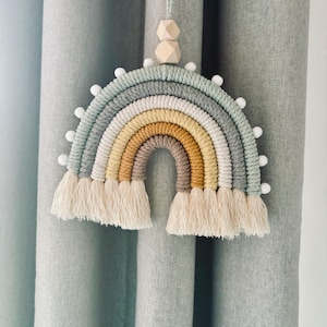 May include: A rainbow macrame wall hanging with a light green, white, yellow, orange, and brown colour scheme. The rainbow has white pom poms around the edge and wooden beads at the top.