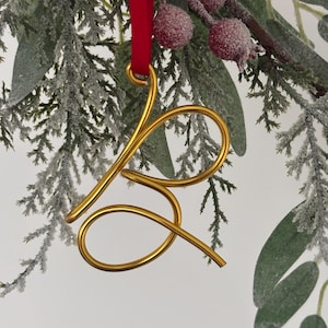 May include: Gold-coloured metal ornament shaped like the letter 'B' hanging from a red ribbon on a snowy evergreen branch.