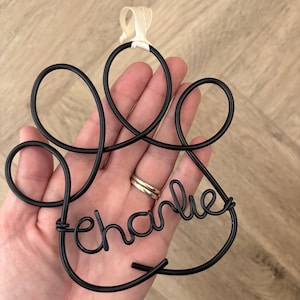 May include: Black wire paw print ornament with the name "Charlie" in cursive script. The ornament is suspended by a cream-coloured ribbon. The design is held in a hand against a light brown wooden surface.
