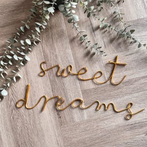 May include: Gold wire script spelling out "sweet dreams" on a wooden surface, with a trailing green plant in the background. The words are in a cursive font, creating a decorative wall art piece.
