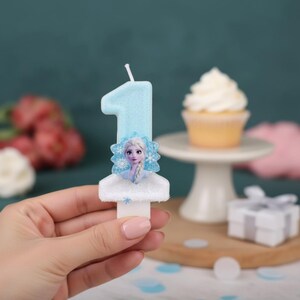 May include: A light blue number one birthday candle featuring a glittery finish and a central graphic of a character with blonde hair and blue clothing. The candle has a white base and a single wick at the top, designed for a cake decoration.