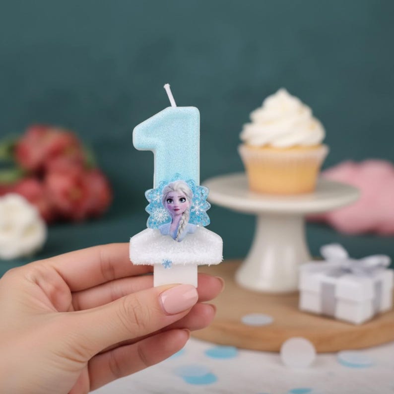 May include: A light blue number one birthday candle featuring a glittery finish and a central graphic of a character with blonde hair and blue clothing. The candle has a white base and a single wick at the top, designed for a cake decoration.