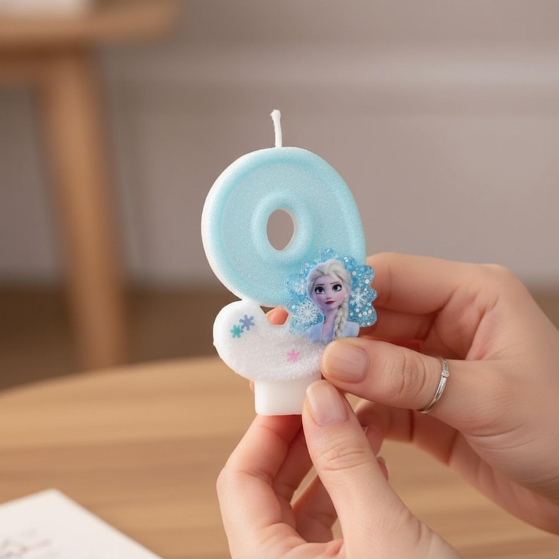 May include: A light blue number nine birthday candle with a glittery finish. The candle features a small, rectangular graphic of a blonde character with a braid, surrounded by blue snowflake accents and a white base with small colored stars.