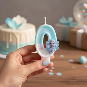 May include: A number zero birthday candle in light blue and white with a textured, glittery finish. The candle features a small, attached graphic of a character with blonde hair and a blue dress, surrounded by blue snowflake accents on the side.