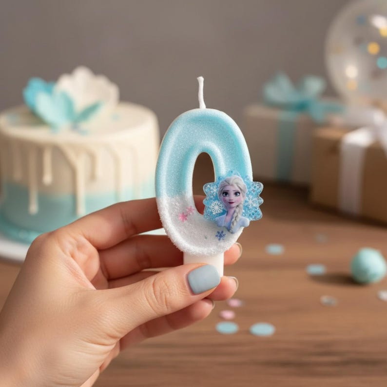 May include: A number zero birthday candle in light blue and white with a textured, glittery finish. The candle features a small, attached graphic of a character with blonde hair and a blue dress, surrounded by blue snowflake accents on the side.