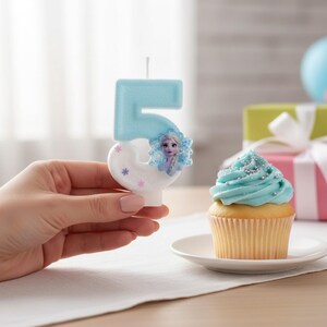 May include: A light blue and white number five birthday candle featuring a small graphic of a character with blonde hair and a snowflake design. The candle has a smooth finish with small purple and pink snowflake accents on the lower curve.