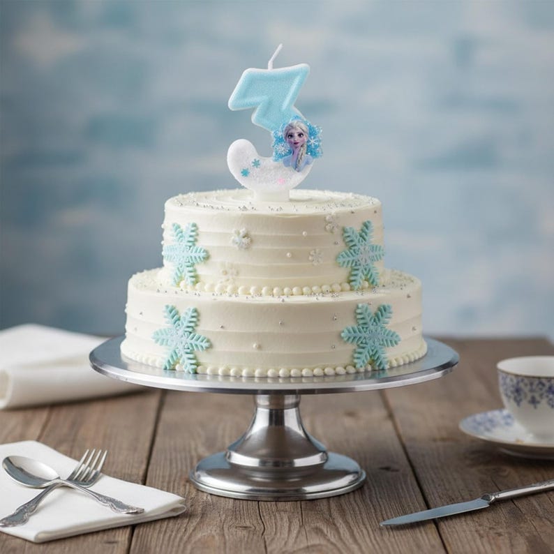 May include: A two-tiered white frosted cake decorated with light blue snowflake accents and silver sprinkles. A number three candle featuring a character illustration sits on top of the cake, which is displayed on a silver metal cake stand.