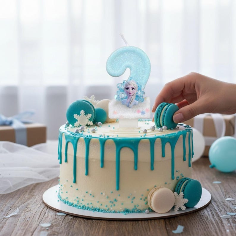 May include: A round cake with white frosting and blue icing drips, decorated with blue and white macarons, silver sprinkles, and white snowflake accents. A glittery light blue number two candle featuring a character image sits on top.