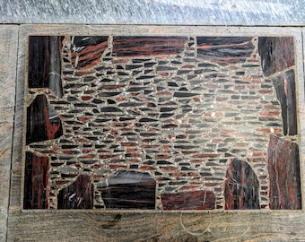 Könnte beinhalten: Ein rechteckiger Steintisch mit einem Mosaikdesign. Das Mosaik zeigt eine Mischung aus roten, schwarzen und grauen Steinstücken, die eine strukturierte Oberfläche bilden. Der Rand ist ein hellerer grauer Stein. Das Gesamtdesign ist abstrakt und künstlerisch.