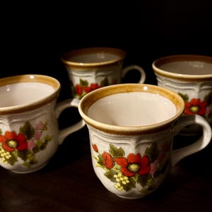 May include: A set of four ceramic mugs with a white base, featuring a floral pattern of red flowers, green leaves, and small yellow clusters. Each mug has a wide, rounded body, a curved handle, and a distinct brown rim around the top edge.