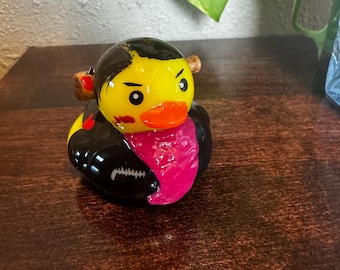 May include: A yellow rubber duck with black hair, a black jacket, and a pink shirt. The duck has an orange beak and is decorated with red accents. The duck is a novelty item, likely a collectible or a decorative piece.