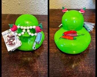 May include: Two green rubber ducks with culinary themes. One duck wears white beaded glasses, a cleaver, and holds a magazine with the text "bon appé". The other duck has chili peppers and a small stack of paper on its body.