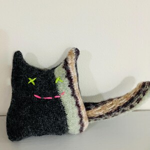 May include: A handmade plush cat toy with a black and white striped body, a black face with green embroidered eyes and a pink stitched mouth, and a long brown tail.