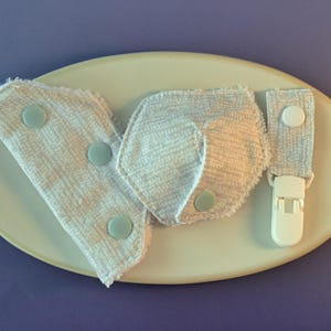 Puede incluir: Tres baberos de tela blanca con ribetes azules y broches. Los baberos están en un plato blanco.