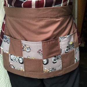 May include: Brown apron with a patchwork design featuring motorcycle images. The apron has a large pocket in the front and a smaller pocket on the side. The text "(c) 2023 - Solstice Days - J Penney" is printed on the apron.