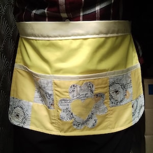 May include: A yellow half apron with a white polka dot waistband and two pockets. The pockets feature a floral design and a bee illustration. The apron is made of fabric.