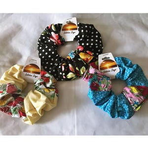 May include: Three fabric scrunchies in various patterns. One is black with white polka dots, another is yellow with floral designs, and the third is blue with floral and geometric patterns. Each has a tag that reads "Solution Days".