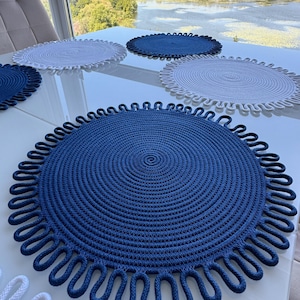 Può includere: Una collezione di tovagliette rotonde nei toni del blu e del bianco. Le tovagliette sono realizzate in corda intrecciata e presentano un motivo a spirale al centro. I bordi sono decorati con un bordo ad anello. Le tovagliette sono disposte su una superficie bianca.