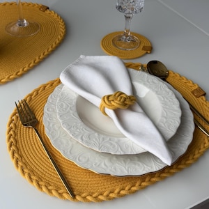 Puede incluir: Una mesa puesta con un salvamanteles, un posavasos y un servilletero trenzados amarillos. Platos blancos con ribetes dorados y una servilleta blanca se disponen con cubiertos dorados y una copa de vino. El servilletero tiene un diseño de nudo.