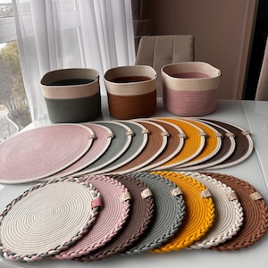May include: A selection of round placemats and matching storage baskets in various colours. The placemats have a braided edge and are in shades of pink, grey, yellow, and brown. The baskets have a two-tone design with neutral and muted colours.