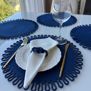 Può includere: Un'apparecchiatura con tovagliette blu navy con bordo smerlato, piatti bianchi, tovaglioli e posate dorate. Sono presenti anche un calice e un portatovagliolo blu navy a forma di nodo.