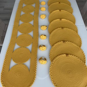 Puede incluir: Un camino de mesa amarillo mostaza que presenta una serie de alfombrillas circulares tejidas conectadas, seis manteles individuales redondos a juego con bordes trenzados y seis servilleteros de nudo, hechos de un material de cuerda grueso.