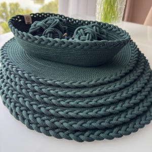 May include: A set of dark green rope items consisting of a shallow oval basket containing two knotted rope spheres, stacked atop a series of circular rope mats. Each piece features a braided edge detail and a small beige fabric tag on the basket.