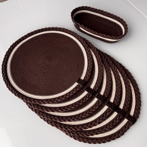 Pode incluir: Um conjunto de seis individuais ovais castanhos com borda trançada e uma faixa creme, juntamente com uma cesta oval combinando. Os individuais têm um design em espiral.