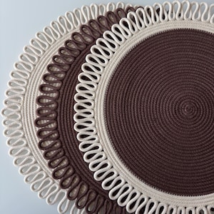 Könnte beinhalten: Zwei kreisförmige Platzsets mit einem gewickelten Seildesign in abwechselnden Bändern aus Dunkelbraun und Creme. Die Außenkanten sind mit einem dekorativen Schlaufenfransenmuster versehen, was ein strukturiertes, geschichtetes Erscheinungsbild erzeugt.