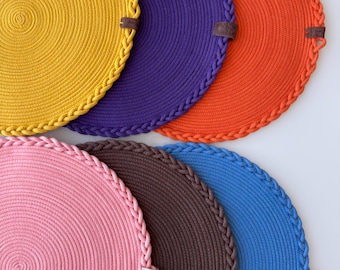 May include: Six round woven placemats in various colors, including yellow, purple, orange, pink, brown, and blue. Each placemat has a braided edge.