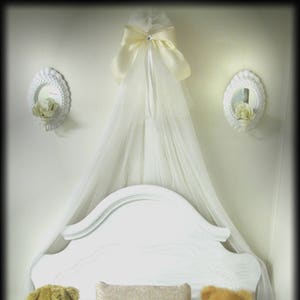 May include: A white canopy bed with a decorative headboard, adorned with a sheer white fabric and a large bow. Three teddy bears in shades of brown and cream are arranged on the bed with a patterned pillow. Two decorative wall sconces flank the bed.