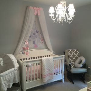 May include: A white wooden crib with a pink and white quilt, positioned beneath a white canopy featuring a pink bow and a castle graphic. A white bassinet stands to the left, while a gray rocking chair with a patterned throw sits to the right.