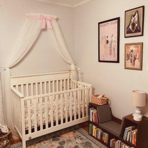 May include: A white crib with a floral patterned mattress is beneath a sheer white canopy with a pink bow. The room features framed vintage art prints, a small bookshelf, and a lamp. A patterned rug covers the floor.