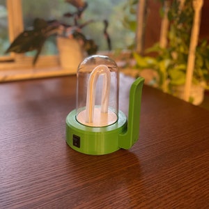 Puede incluir: Una lámpara de crecimiento para plantas verde y transparente con una bombilla fluorescente curva dentro de una cúpula de cristal. La lámpara tiene una base verde con un interruptor de encendido/apagado y un asa. La lámpara está sobre una superficie de madera marrón.