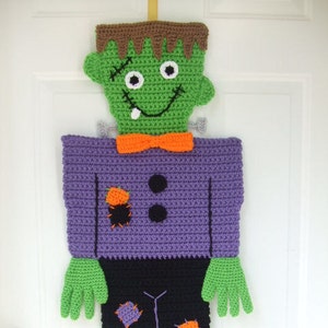 Può includere: Una decorazione di Halloween all'uncinetto di un mostro di Frankenstein verde con una camicia viola, pantaloni neri e scarpe marroni. Il mostro ha una cicatrice cucita sul viso e un ghigno pieno di denti.