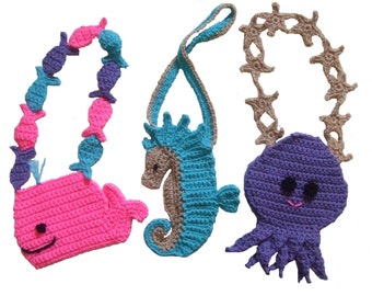 May include: Three crocheted sea creature purses with straps. The first is a pink whale with a blue and purple fish strap. The second is a brown and blue seahorse with a brown strap. The third is a purple octopus with a brown starfish strap.