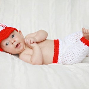 May include: A baby wearing a white and red crochet baseball cap and matching pants. The baby is lying on a white textured blanket. The hat and pants have a red trim.