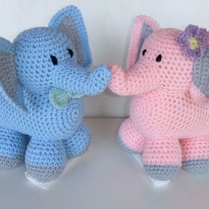 Può includere: Due giocattoli di elefante all'uncinetto, uno blu e uno rosa, con orecchie e piedi grigi. L'elefante rosa ha un fiore viola sulla testa.