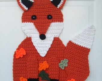 May include: A crocheted fox door hanger with a white tail and orange body. The fox has black eyes and a black nose. The fox is surrounded by green and orange leaves.