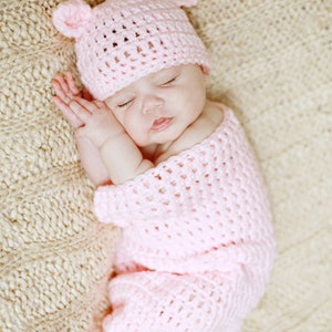 Può includere: Un bambino che dorme con un cappello e un completo di uncinetto rosa. Il bambino è sdraiato su una coperta di maglia beige.