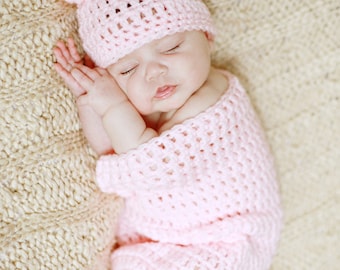 May include: A sleeping baby wearing a pink crocheted hat and matching crocheted outfit. The baby is lying on a beige knitted blanket.