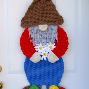 Puede incluir: Un colgador de puerta de gnomo de ganchillo con un sombrero marrón, una camisa roja, pantalones azules, barba blanca y flores blancas. El gnomo está de pie sobre una base marrón con setas verdes, amarillas y rojas.