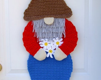 May include: A crocheted gnome door hanger with a brown hat, red shirt, blue pants, white beard, and white flowers. The gnome is standing on a brown base with green, yellow, and red mushrooms.