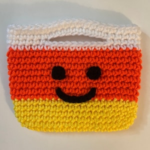 Puede incluir: Una bolsa de ganchillo con forma de maíz dulce con un asa blanca y una cara sonriente. La bolsa es naranja, amarilla y blanca.