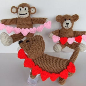 May include: Three crocheted stuffed animals wearing pink and red heart garlands. A brown monkey, a brown bear, and a brown dog.