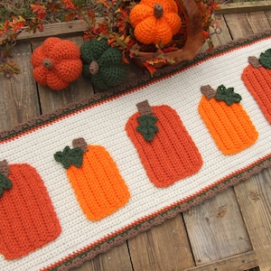 May include: A crocheted pumpkin runner with a white background and brown and green trim. The runner features five orange pumpkins with green leaves.