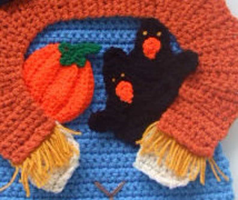 May include: A blue crocheted sweater with a pumpkin, ghost, and orange scarf. The sweater has a brown stitched design on the front.