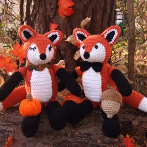 May include: Two crocheted fox plushies sitting on a log. The fox on the left is holding a small orange pumpkin and the fox on the right is holding a brown acorn. Both foxes have black eyes, white fur, and orange fur.